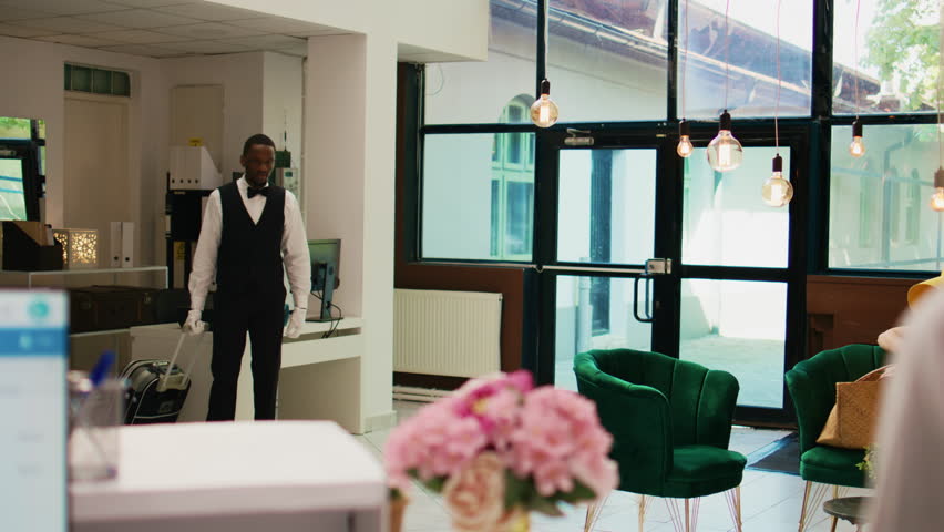 Hotel bellhop carrying lugagge in lobby, providing luxury service to hotel guests arriving on tropical summer vacation. African american employee helping people with trolley bags, bellboy.