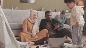Slowmo of happy interracial Muslim couple and their little son video chatting with friends or relatives on laptop from home, smiling and waving at web camera