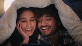 Multiracial happy carefree couple Indian man Hispanic woman watching film movie funny video laughing smiling having fun humorous ridiculous news joke mobile phone laugh cover under blanket at night - Powered by Shutterstock - Get 15% off with code: PIKWIZARD15