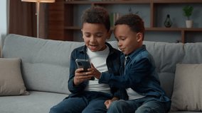 Two little African American siblings boys sitting on couch using mobile phone talking scrolling smartphone play online game. Ethnic children kids brothers browsing cellphone app together talk at home