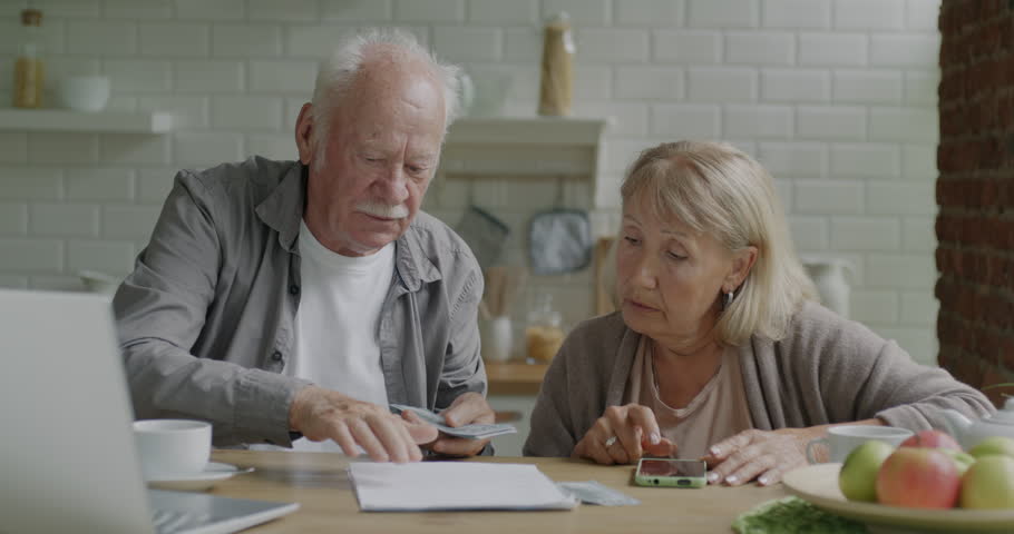Unhappy senior people counting money paying expensive bills checking payment documents in kitchen at home. Family budget and poverty concept.