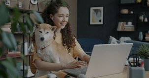 Young woman hugging shiba inu dog and making online video call with laptop and wireless earphones at desk at home. Communication and modern technology concept.