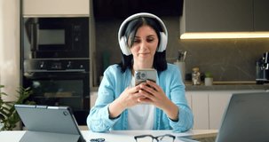 Modern, happy and relaxed woman with headphones holding mobile phone watching videos and listening to favorite music podcast or online social media at home office.