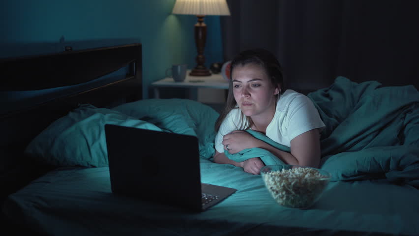 Happy young woman eating popcorn while lying on bed watching movie on laptop at night at home Royalty-Free Stock Footage #1105100041
