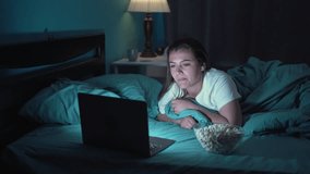Happy young woman eating popcorn while lying on bed watching movie on laptop at night at home - Powered by Shutterstock - Get 15% off with code: PIKWIZARD15