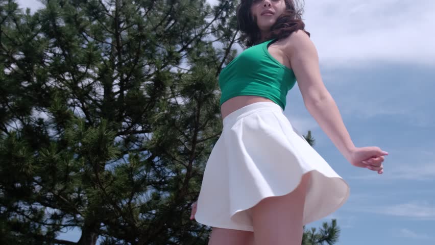 Beautiful girl in a green top and white shorts is spinning and having fun in a city park against the backdrop of a coniferous tree. Slow motion

