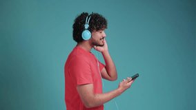Cheerful handsome young swarthy guy in casual watching videos on smartphone, using new headset, looking at cell phone and smiling, in t-shirt on blue studio background, copy space