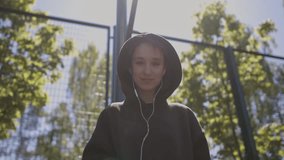 A smiling girl in a sweatshirt on the sports ground listens to music on headphones, holds a smartphone in her hands and looks at the camera. Girl with headphones and smartphone, looks at the camera.