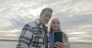 Elderly couple walking near river. Stylish mature lovers smiling and looking at smartphone. Senior married couple have video chat with children. Woman holding gadget and talking,