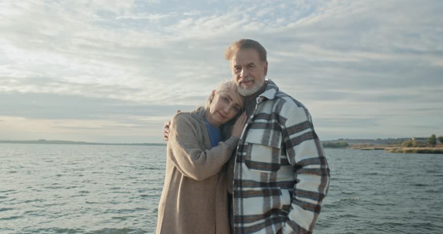Senior married couple smiling and posing for camera. Mature couple in love at background of lake and sky. Husband hold wife tightly. Bearded man and preety woman enjoy being together.
