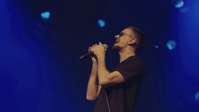 Professional male singer with microphone in dark glasses and black shirt performs on stage. Blue and red spotlights. Slow motion