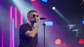 Young attractive man in black glasses sings into microphone on stage of nightclub. Slow motion