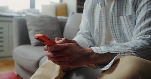A man in casual clothes smiles while typing on a mobile phone while sitting on the couch at home. The video moves from the man's hands to his face