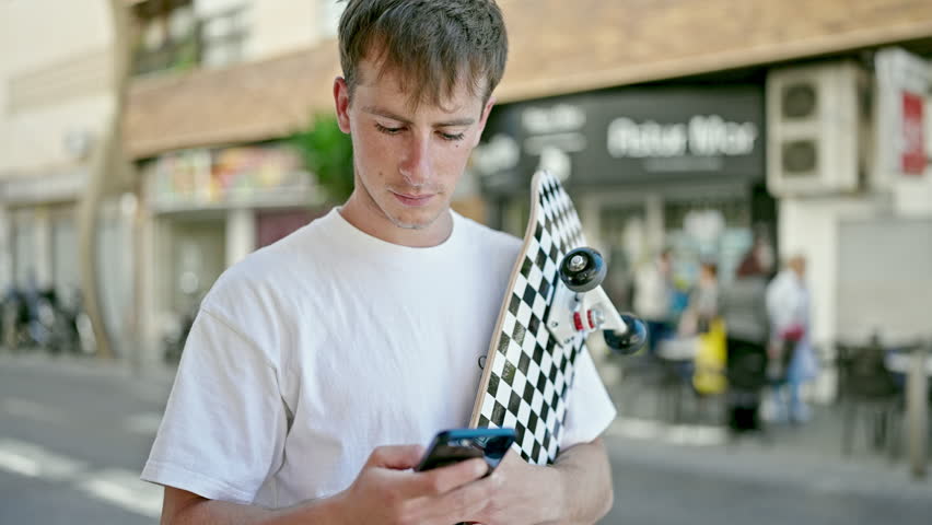 Young caucasian man using smartphone holding skate at street