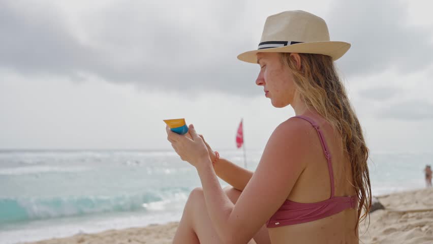 A young woman in a pink swimsuit, holding sunscreen cream in her hand.