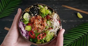 Person holding a poke bowl, showing poke with raw salmon and fresh vegetables and rice served in wooden plate on tropical background, top view video clip, close up 4k footage