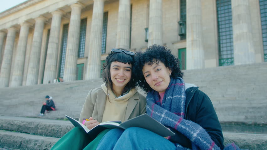 Two young cheerful female students sitting with copybooks on staircase in front of university, looking at camera and smiling. Video portrait, handheld shot