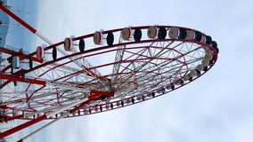 Red and white ferris wheel is rotating against grey cloudy sky at amusement park - vertical video. Sightseeing, cityscape, entertainment and holiday concept