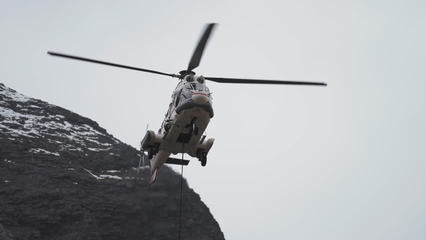 Cargo helicopter with lifting cable and load attached landing in the difficult-to-reach mountainous terrain. Snow-covered mountains in the background. View from below. Pan follow.