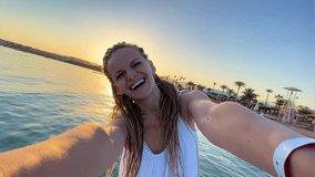 Happy smiling girl joyfully making excited selfie video call in summer vacation, young woman with dreadlocks taking selfies over sunset ocean sea beach, slow motion