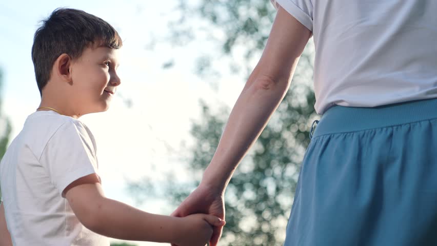 Happy kid walks through park with green trees holding her mom hand and smiling. Mom son, holding hand, walking in park on summer day. Concept family relations. Happy family relaxing together in nature
