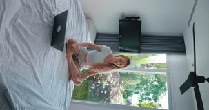 Vertical video pretty blonde woman sitting on bed with laptop and meditating breathing raising arms for stress relief, relaxing from work. Millennial female practices yoga meditating at home bedroom