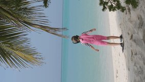 Vertical video. Rear view of little girl going through the tropical beach to snorkeling in ocean water, slow motion. Summer vacation and travel lifestyle concept
