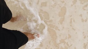 VERTICAL VIDEO: Man wearing shorts, walking barefoot along the seashore. Male legs walks on sandy beach along the shore near the water with waves, top view, POV. Travel in summertime concept.