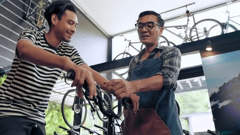 Asian senior salesman teaching use brake of bike to customer young man buying new bike in shop store. Small business and dealer concept วิดีโอสต็อก