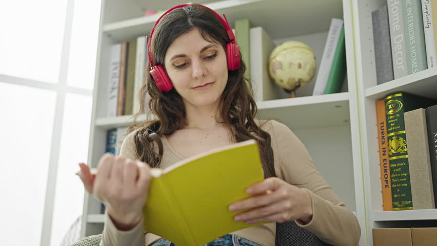 Young beautiful hispanic woman student listening to music reading book at library university