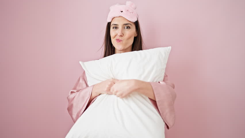 Young beautiful hispanic woman wearing sleep mask hugging pillow smiling over isolated pink background