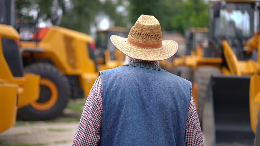 Back view tracking shot of experienced senior farmer looking around walking between rows of new yellow tractors. Live camera follows confident professional Caucasian man choosing new vehicle Royalty-Free Stock Footage #1105183967