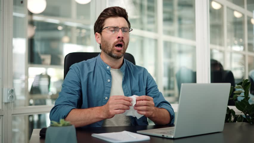 Unhealthy male employee worker sneezing while working in workplace in office. Sick young businessman working on computer pc using napkins. Tired ill managers sitting at desktop, feeling bad, cold. Royalty-Free Stock Footage #1105188471
