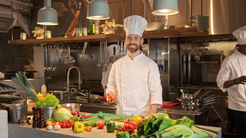 Portrait of male chef standing and juggles tomato at the background of work process at the restaurant's kitchen. Proffesional group of multicultural cooks in special uniforms working together.