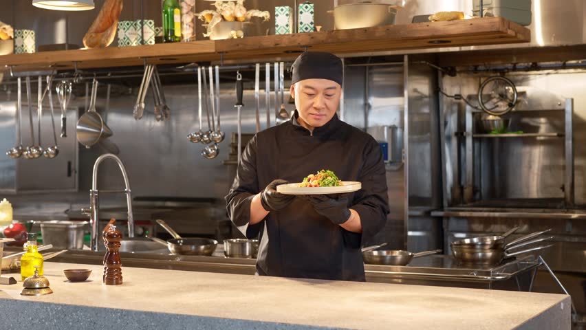 Asian man in special blac uniform stands in restaurant professional kitchen. Chef holding plate with vegetables salad, looking at camera and smiling on background of empty kitchen.