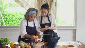 happy asian young daughter and senior mother cooking online class on tablet together making fresh vegetables food in kitchen at home