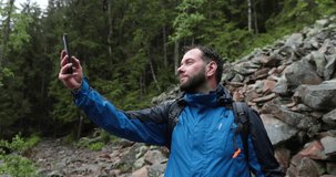 A man stands in the forest on large stones and takes out a smartphone to search for a network and make a call. Hiking in the mountains in rainy weather. He has a backpack on his shoulders.