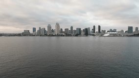 Drone aerial view of Southern California city and skyline of San Diego on a cloudy summer day moving towards downtown skyscrapers and buildings from above the bay water surface with boats in harbor