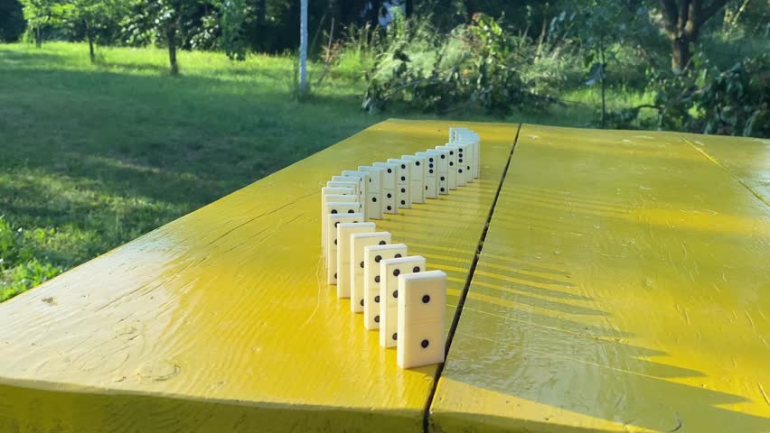 Dominoes falling on yellow table by touching of female hand. Cause and effect concept. Domino effect. Beautiful landscape garden on background. Chain reaction as analogy to sets events caused by one
