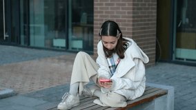 A young girl schoolgirl sits on a bench and reads messages in her smartphone