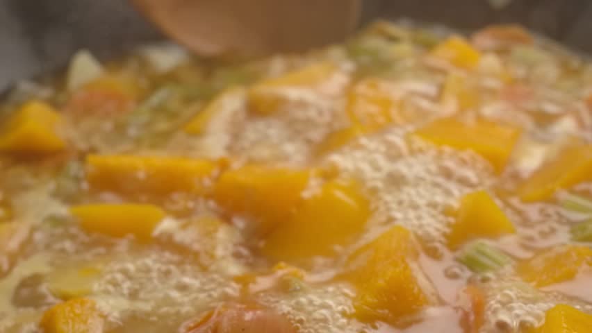 Close-up of the wooden spoon mixing pumpkin soup boiling with the vegetables in the pan in slow motion