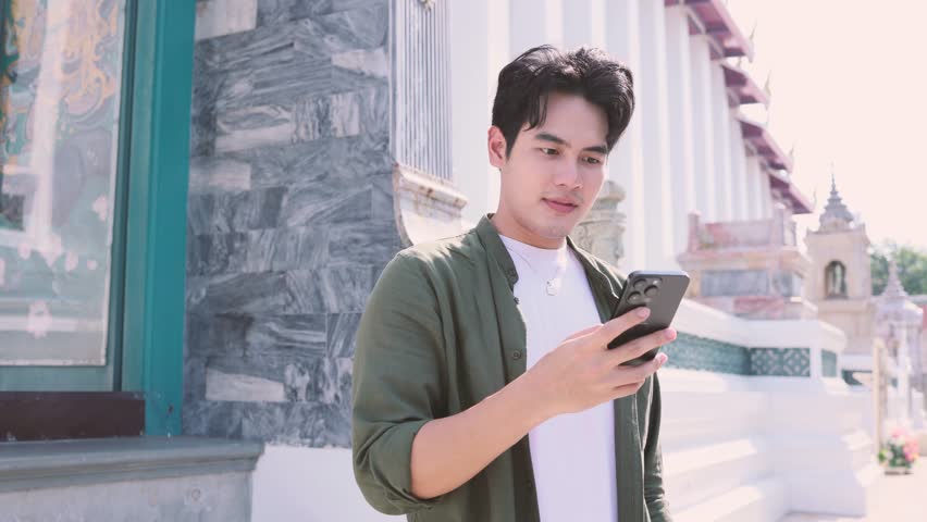 Portrait of Asian male traveler using a mobile phone on sidewalk of buddhist temple on street in Bangkok, Thailand, Southeast Asia - smart phone and internet for traveling concept Royalty-Free Stock Footage #1105217743