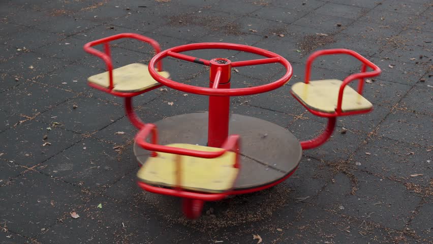 An empty red children carousel spins in a city park after rain close up. Lonely swing is spinning on the playground in the yard. Old deserted playing field without children. Video footage in 4k 25FPS Royalty-Free Stock Footage #1105219509