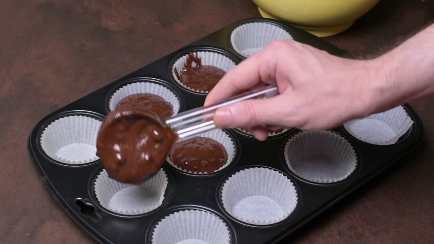 Portioning of chocolate muffin batter into paper molds in muffin tray. Male hand. Close-up. Royalty-Free Stock Footage #1105220487