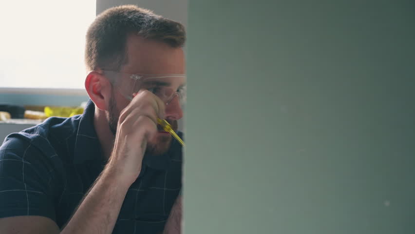 CU, Dolly cam: nervous young man in protective goggles and blue shirt repairs plug on wall with screwdriver in light room Royalty-Free Stock Footage #1105221201