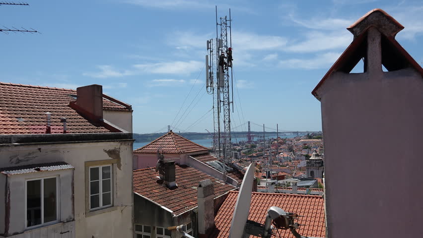 Antenna Workers Tower Risky Job Building Tops City Skyline. Male workers fixing an antenna hanging on top of the tower above city building roofs