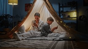 Full length portrait of a multi ethnic young woman, loving caring mother sitting with her child on floor inside a play tent wigwam, having fun together, playing online video games. People. Technology