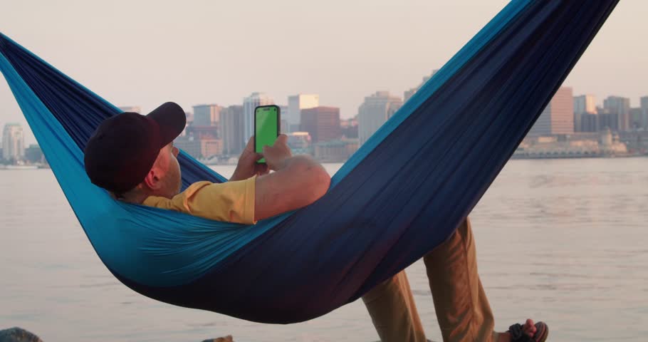 Man in a Hammock Holds a Mobile Phone Vertically with His Hand and Swipes His Finger Across the Touch Screen. Use Green Screen for Copy Space. Chroma Key Mockup on Smartphone.