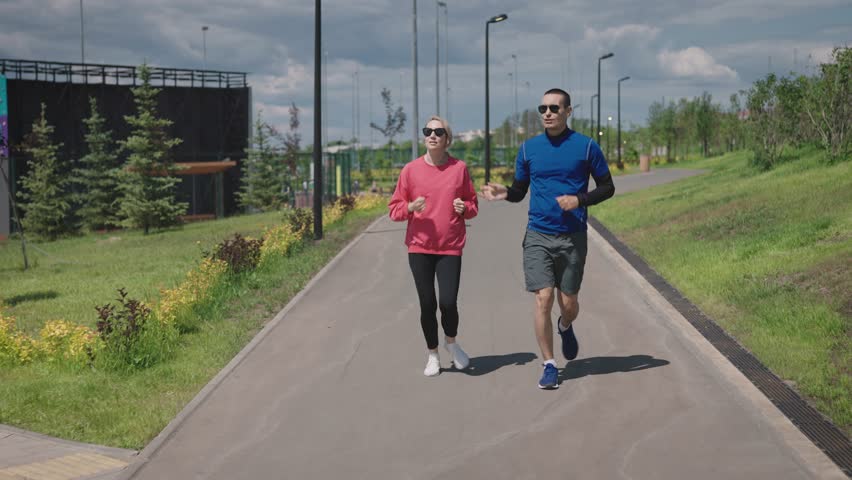 Sports couple on a daily run in the city park. Bright summer day and landscaped park area. Convenient urban environment for sports and recreation. Royalty-Free Stock Footage #1105230295