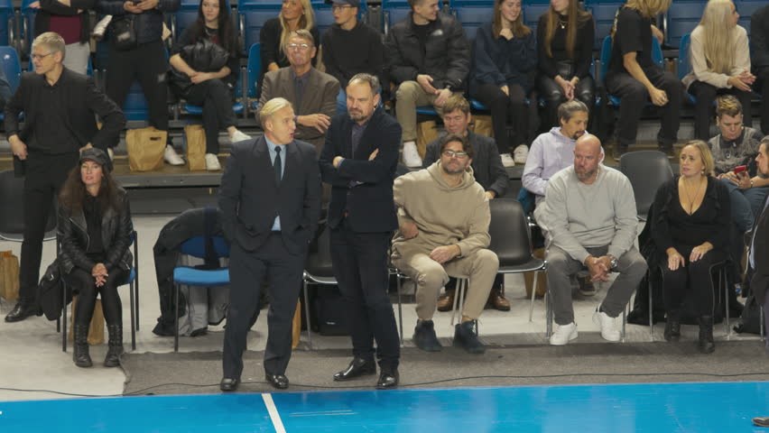 Group of Basketball Coaches and Managers Wearing Suits, Discussing In-Game Strategy Next to The Court with Fans in the Background. Trainers Discussing Offensive and Defensive Team Conditioning